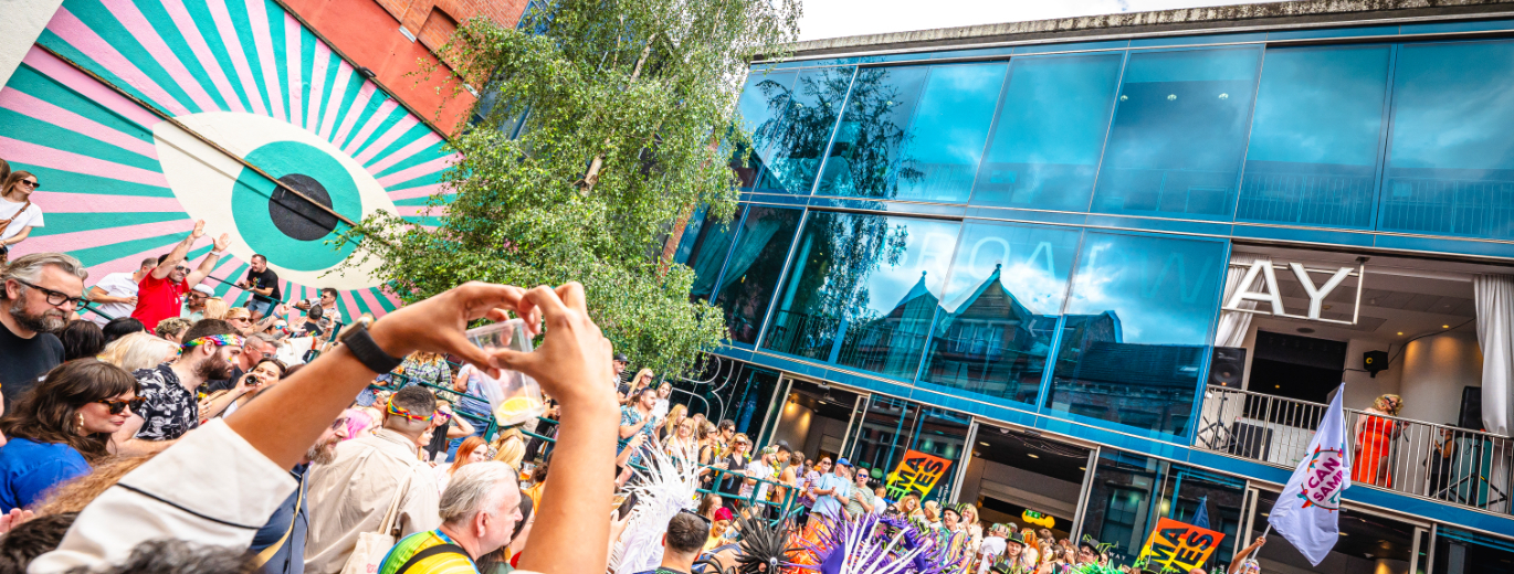 A vibrant street scene with a crowd of people gathered for an event. In the foreground, a person forms a heart shape with their hands, framing the view. The background features a large building with a colourful mural on an adjacent building with geometric patterns and an eye design. The atmosphere appears festive, indicated by flags and banners held by individuals in the crowd.