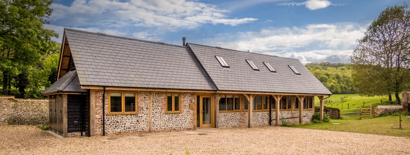 A small brick building with a slate roof and wooden beams from the roof to the floor at the front with lots of greenery behind.
