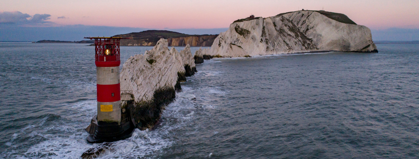 "White cliffs with the sea crashing against them, with a red and white lighthouse at the end of a row of rocks"