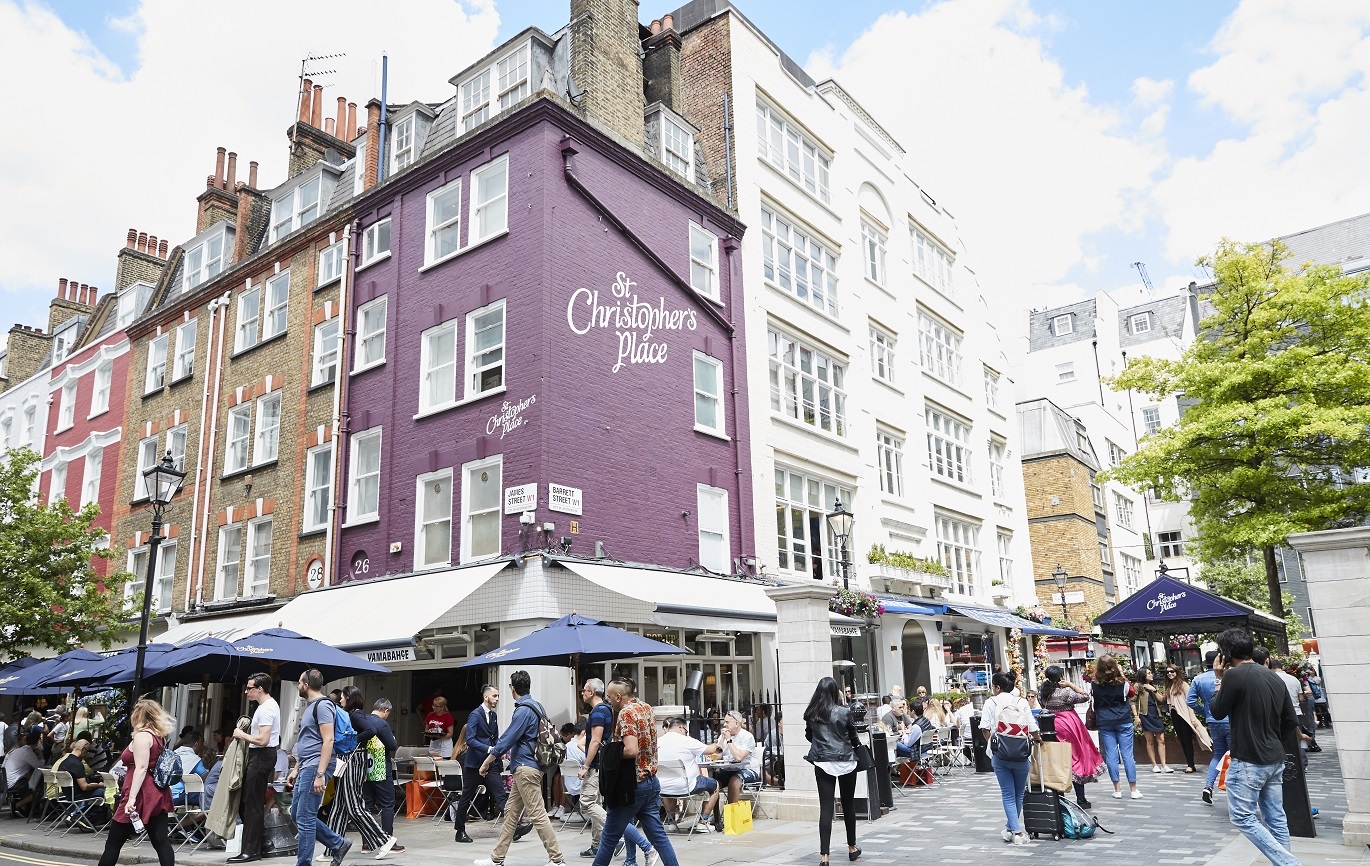"Photo of St Christopher's Place written in white on a purple wall of a building on the corner in the area. There is a restaurant below with people walking around the area."