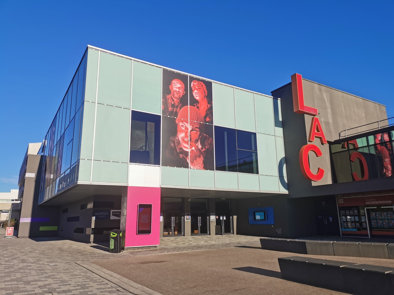 "Photo of the frontage of Lincoln Arts centre with the entrance and graphics on the wall, Lincoln Arts Centre is written on the wall and it says Learn, Make, Watch, Debate above the entrance. There are blue skies"