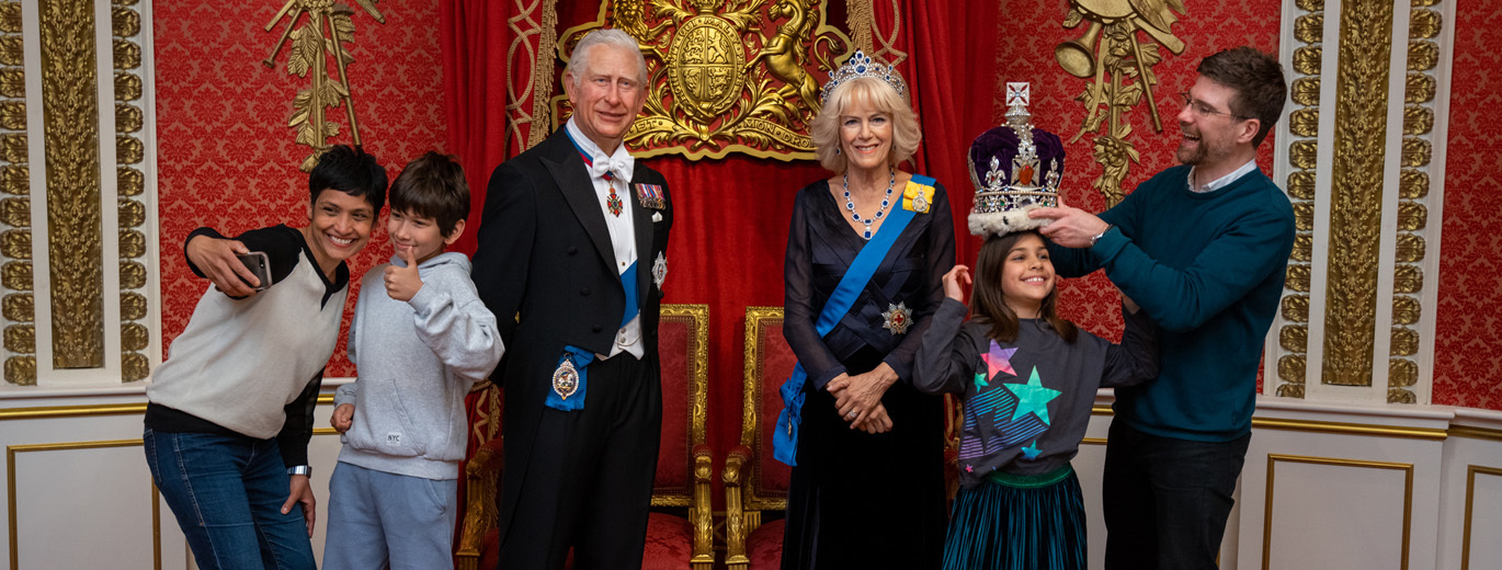 "A group of people posing with wax figures of King Charles and Queen Camilla stood in front of a red and gold throne."