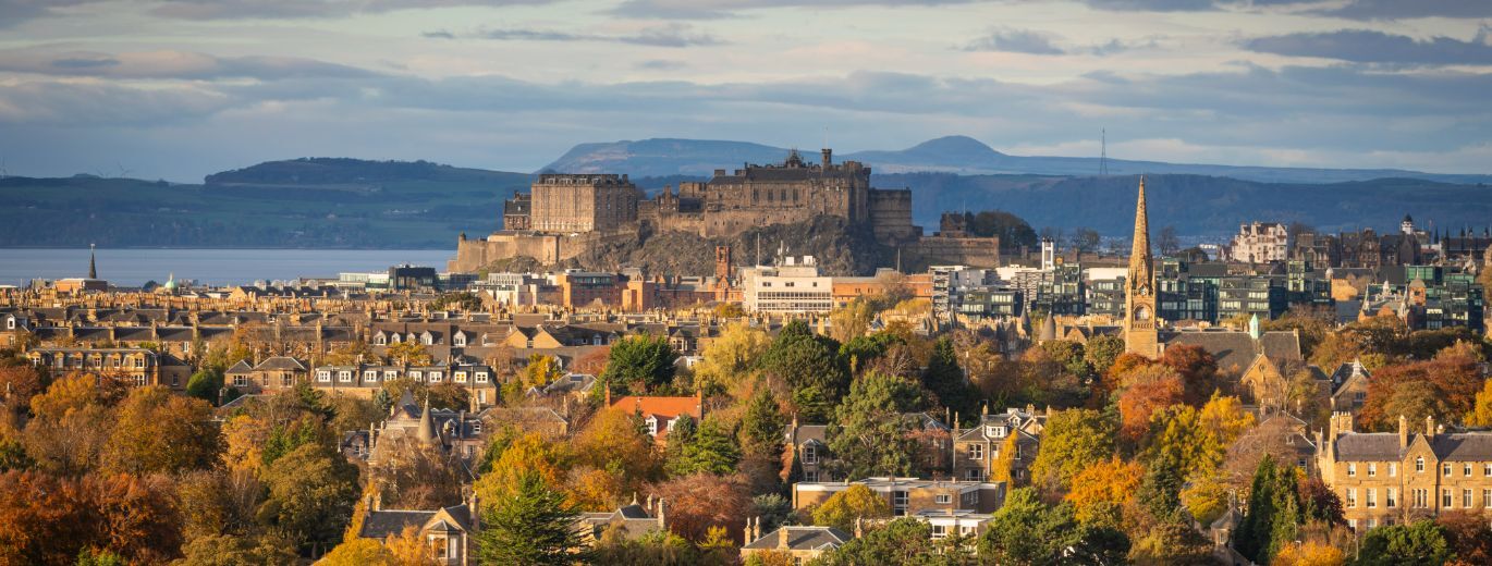 "Spectacular view of Edinburgh and Edinburgh castle from a distance. The foreground is trees and homes, and the castle is rising above the city in the middle. In the distance are rolling hills"