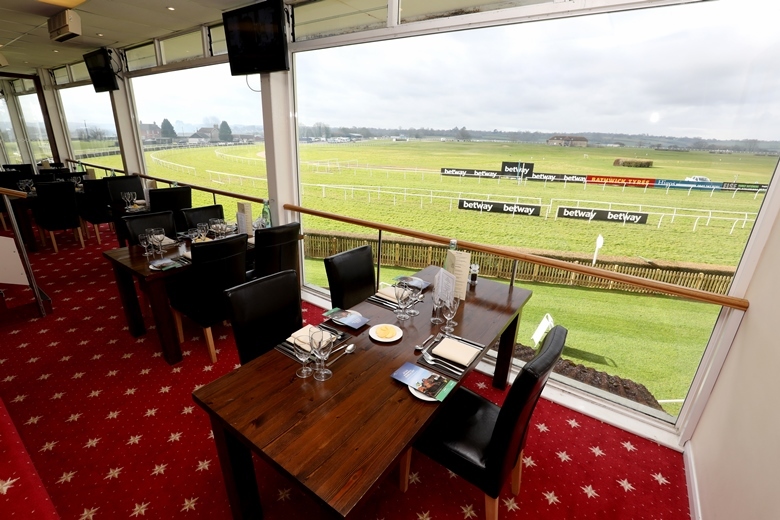Restaurant-style seating area overlooking a horse racing track through large floor-to-ceiling windows, with tables set on a red patterned carpet and the racecourse visible outside.