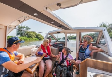 People seated inside a motorboat with a glass canopy, conversing as the boat moves along a river bordered by green trees.