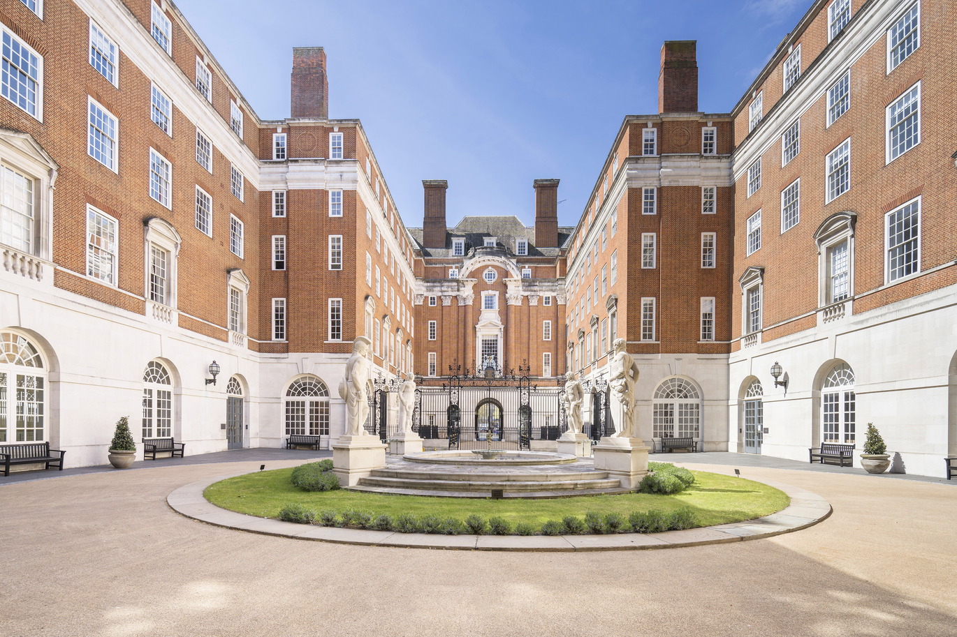 Courtyard of a large red-brick building with white stone ground floor, arranged around a circular garden with a central fountain, under a clear blue sky.