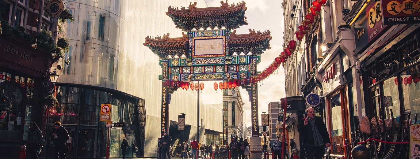 "Photo of lots of people walking through Chinatown, there are restaurants either side of the path and a large archway decorated in lanterns and symbols"