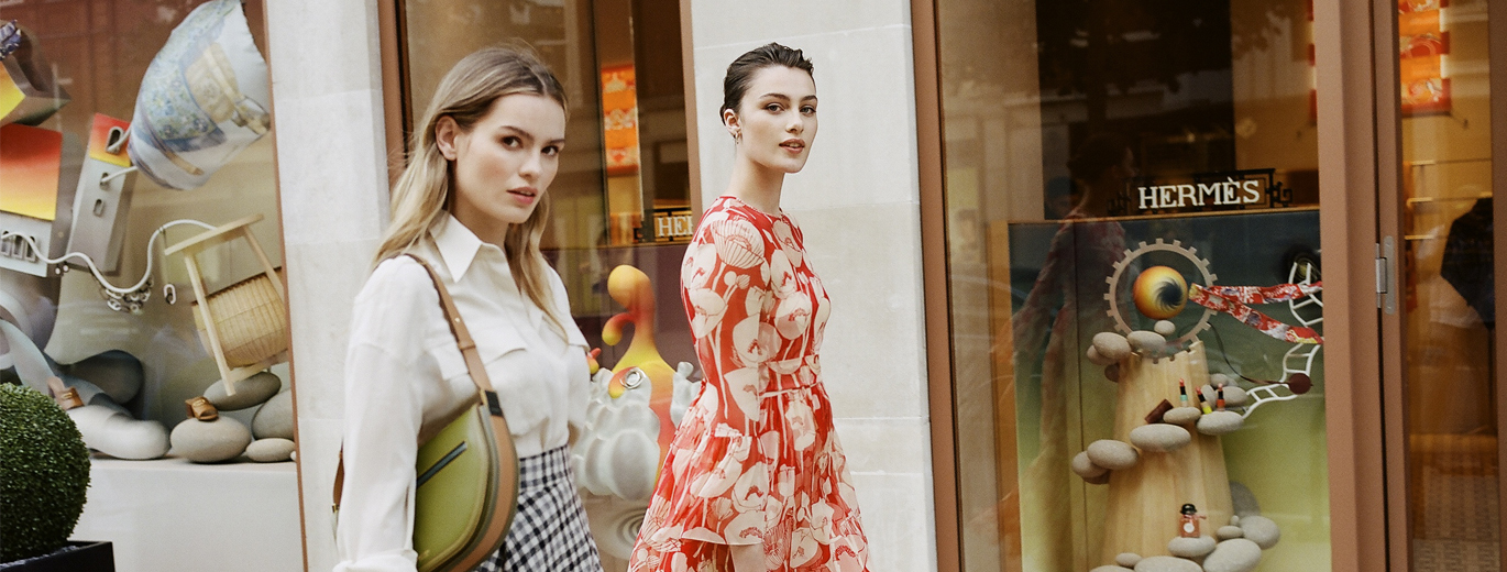 Photo of 2 people looking at the camera while walking past a Hermes shop. The person on the left is wearing a white shirt and checkered skirt holding a green bag. The person on the right s in a red and white dress.