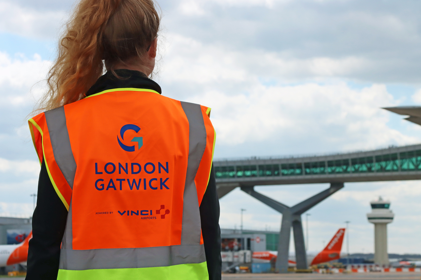 "Person with long hair in a ponytail and neon orange jacket with London Gatwick name and logo on the back, facing a London Gatwick building"