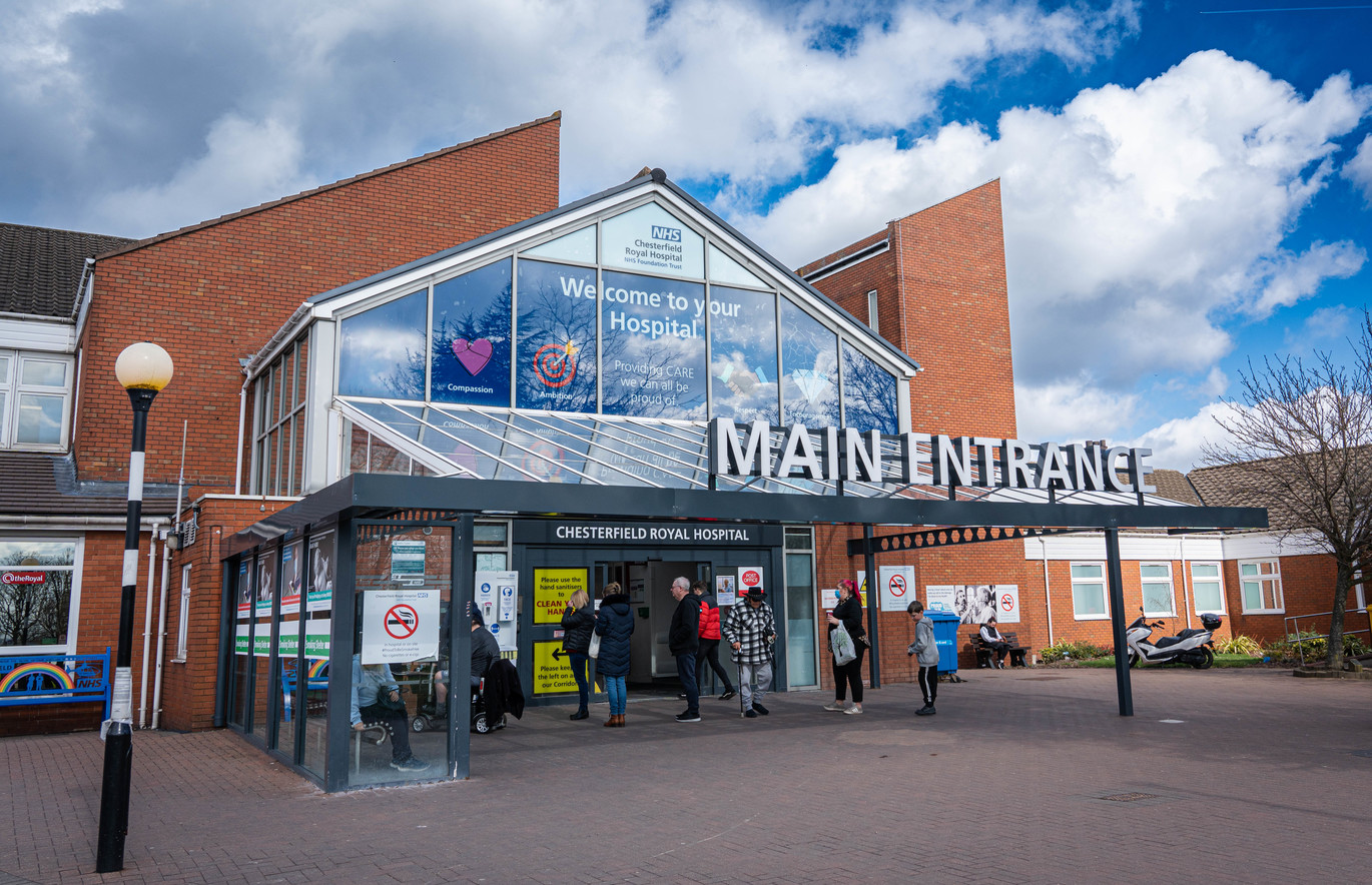 Front entrance of a hospital building with a glass facade and large “MAIN ENTRANCE” sign, people walking in and out; separate image of the NHS logo in white on a blue background.