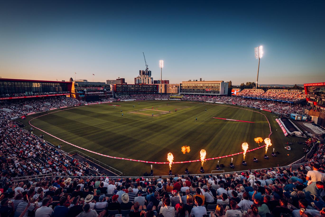 Old Trafford stadium getting ready for a day night match, fire going off in the foreground, the batters are entering the pitch
