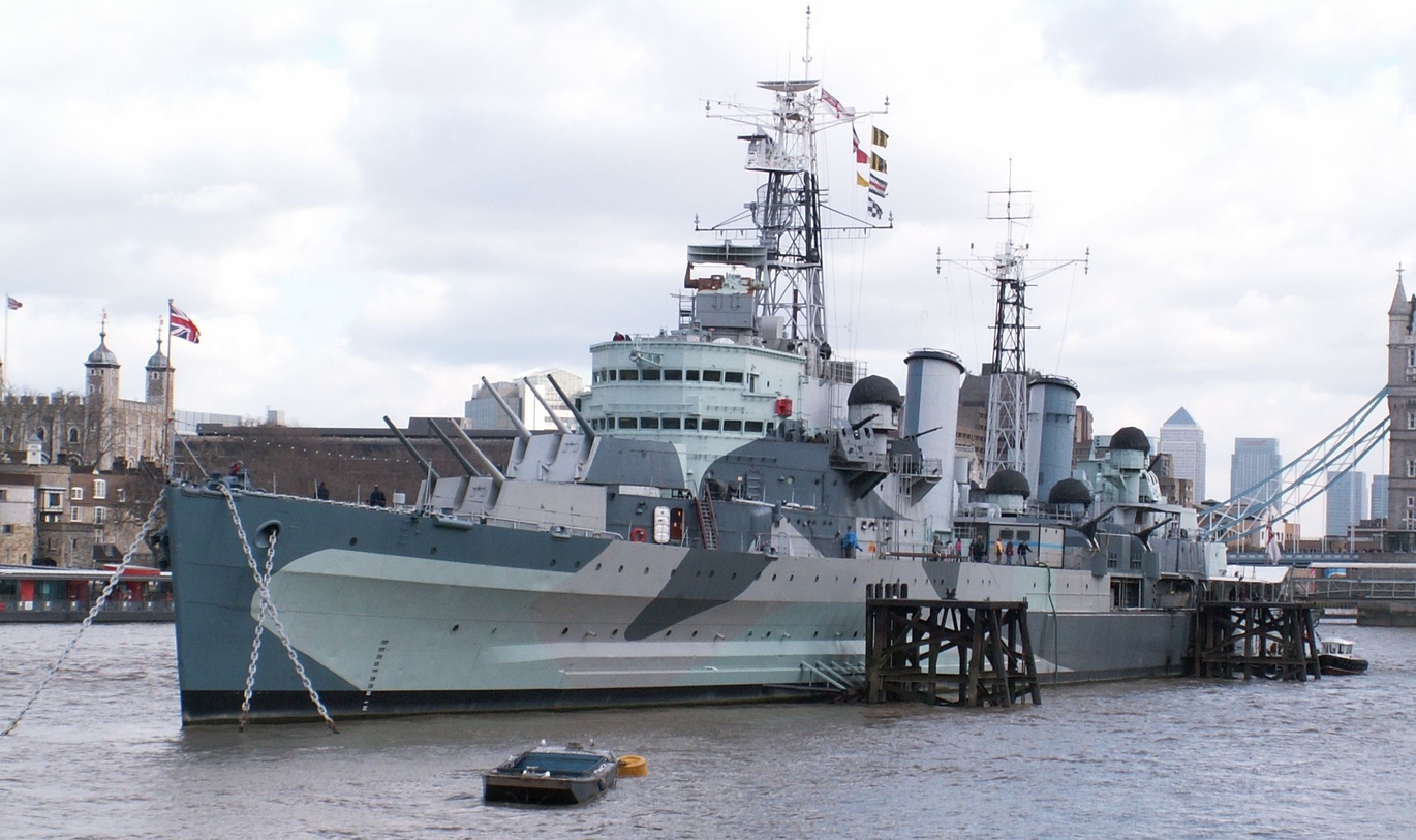 Image of HMS Belfast docked. The ship is shades of blue, green and grey. There are flags flying at the top. There is a view of the local London area in the background.