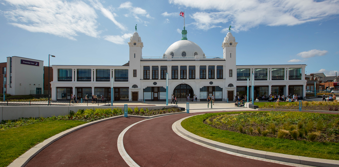 A large white building with a central dome and small towers, featuring an arched entrance and rows of windows, sits behind a curved driveway and landscaped greenery under a blue sky.