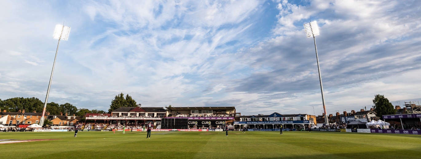 Game of cricket taking place at County ground, sun out, whites on.