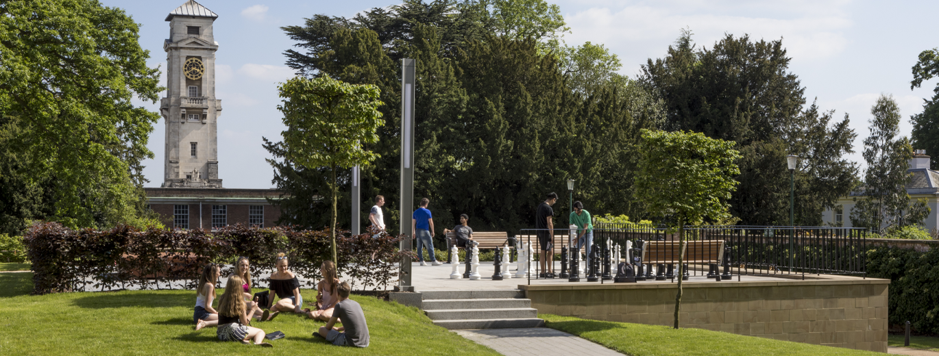 "Students enjoying a sunny day sitting on the grass, in the background is a giant chess board, the tower of Trent building can be seen in the background"