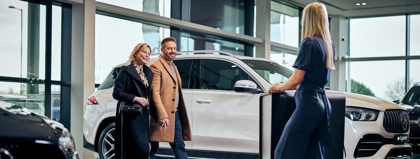 "A colleague approaches a couple in a Mercedes-Benz dealership, with a white SUV in the background."
