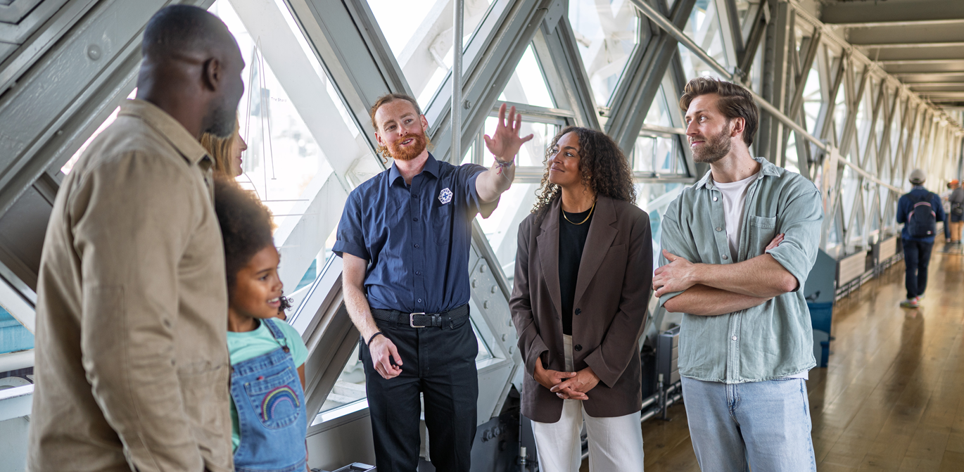 "A Tower Bridge colleague showing 4 adults and 1 child the area on an enclosed bridge area with glass walls"