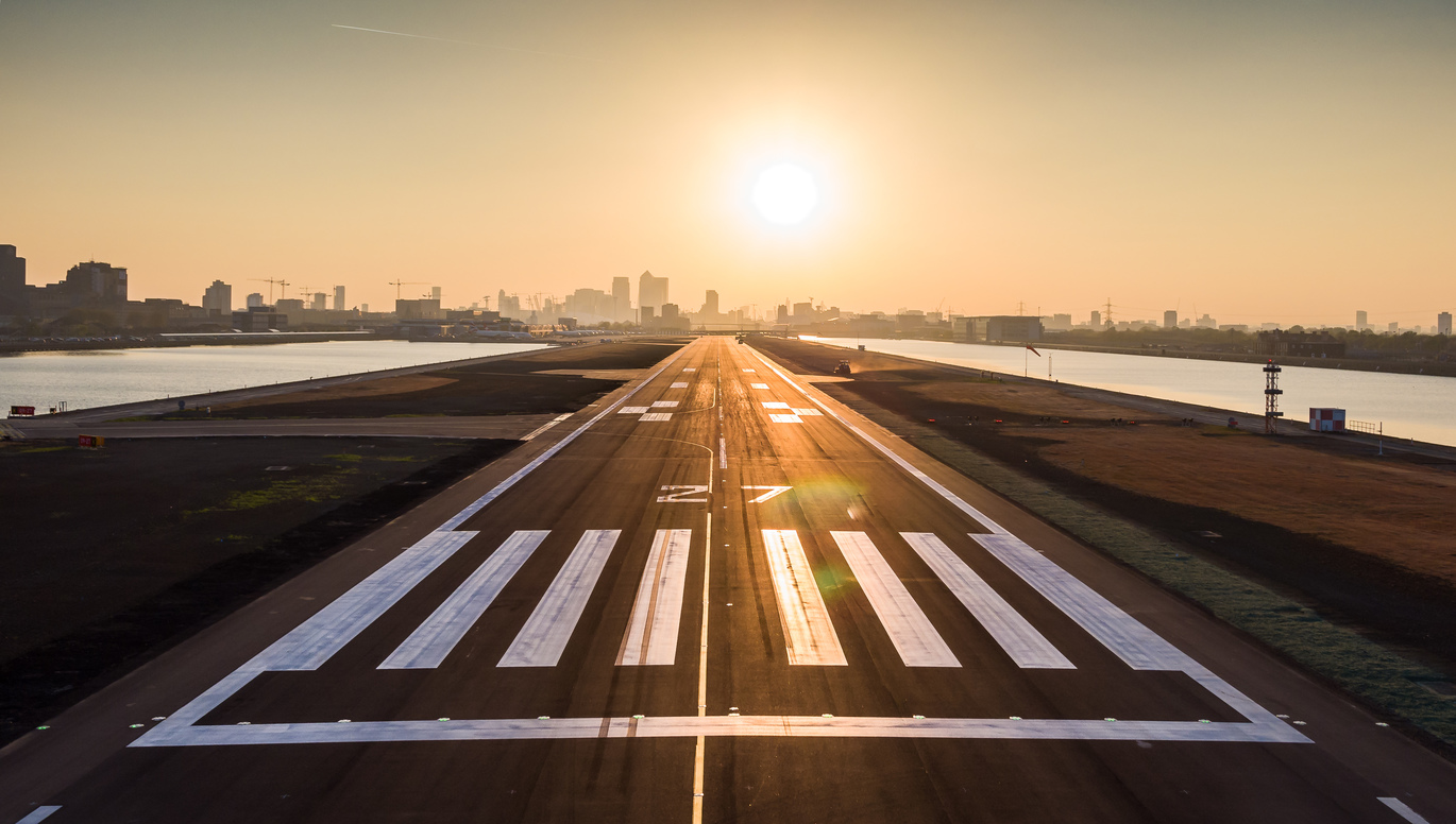 "London City Airport Runway with the city skyline in the background and the sun directly in the middle shining on the runway"