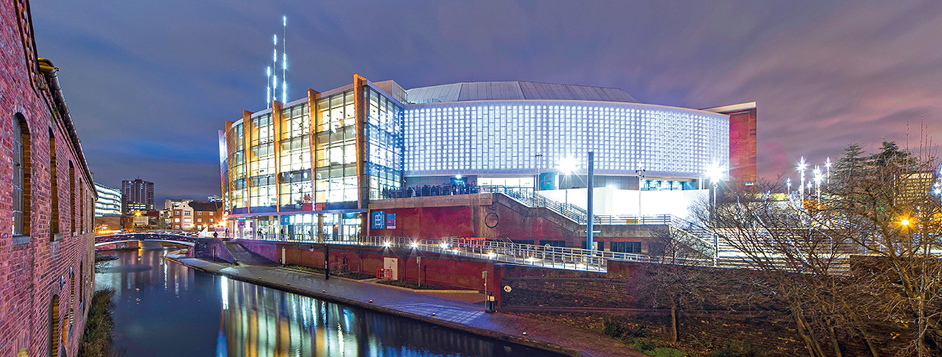 "A photo of Utilita Arena Birmingham exterior from across the river. The river is at the front of the photo with the venue in the background, there is a bridge across the river to the left of the photo. The photo is taken at night and the venue is lit up throughout."