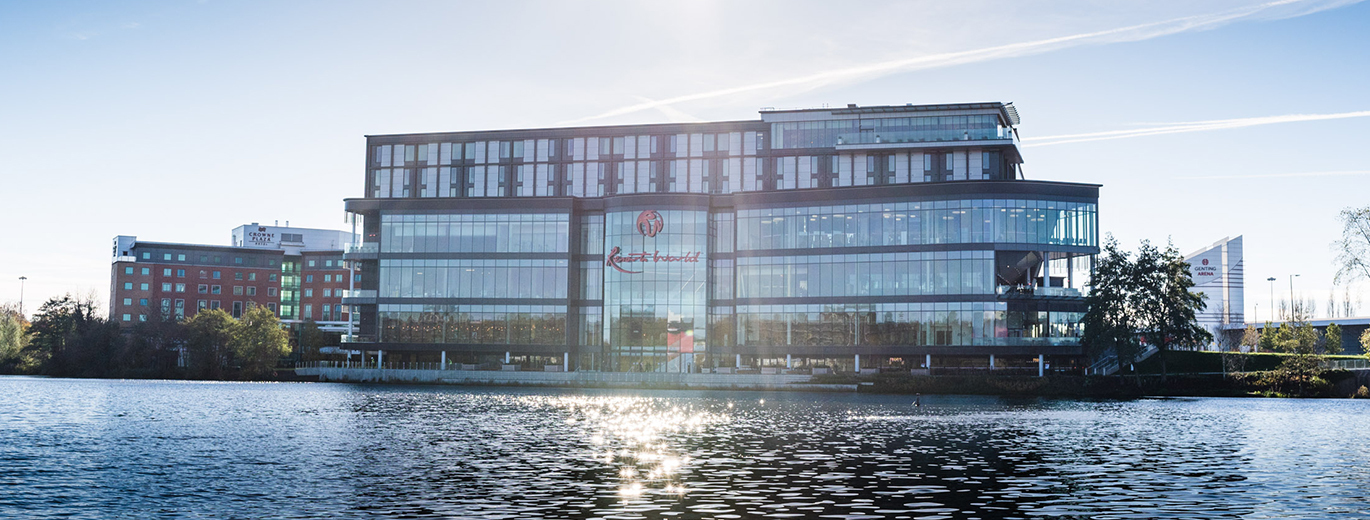"Photo of Resorts World exterior from across the river. The river is at the front of the photo with the Shopping Centre in the distance. There is a Resorts World logo in the middle at the top of the building."
