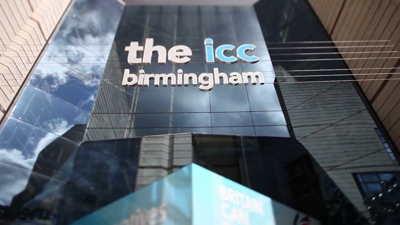 Exterior view of a glass-fronted building with the words “the icc birmingham” displayed in white and blue lettering across the façade, reflecting the sky and surrounding structures.