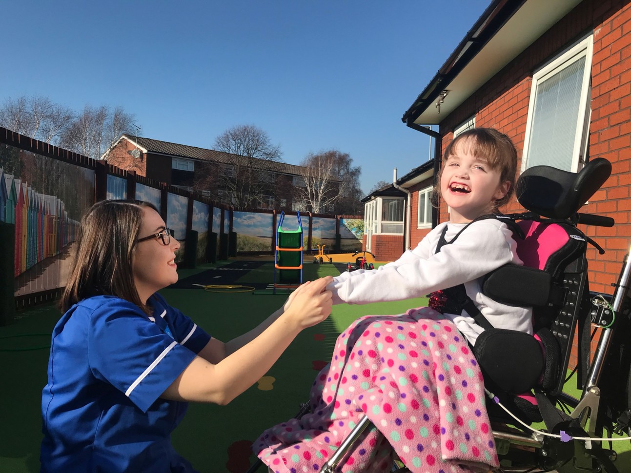 Child in a powerchair is smiling at the camera as she holds the hands of a nurse who smiles at her