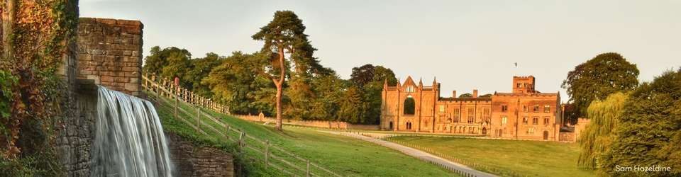 Historic estate with a stone waterfall in the foreground, a grassy park and trees, and a large gothic-style manor glowing in warm evening light in the background.