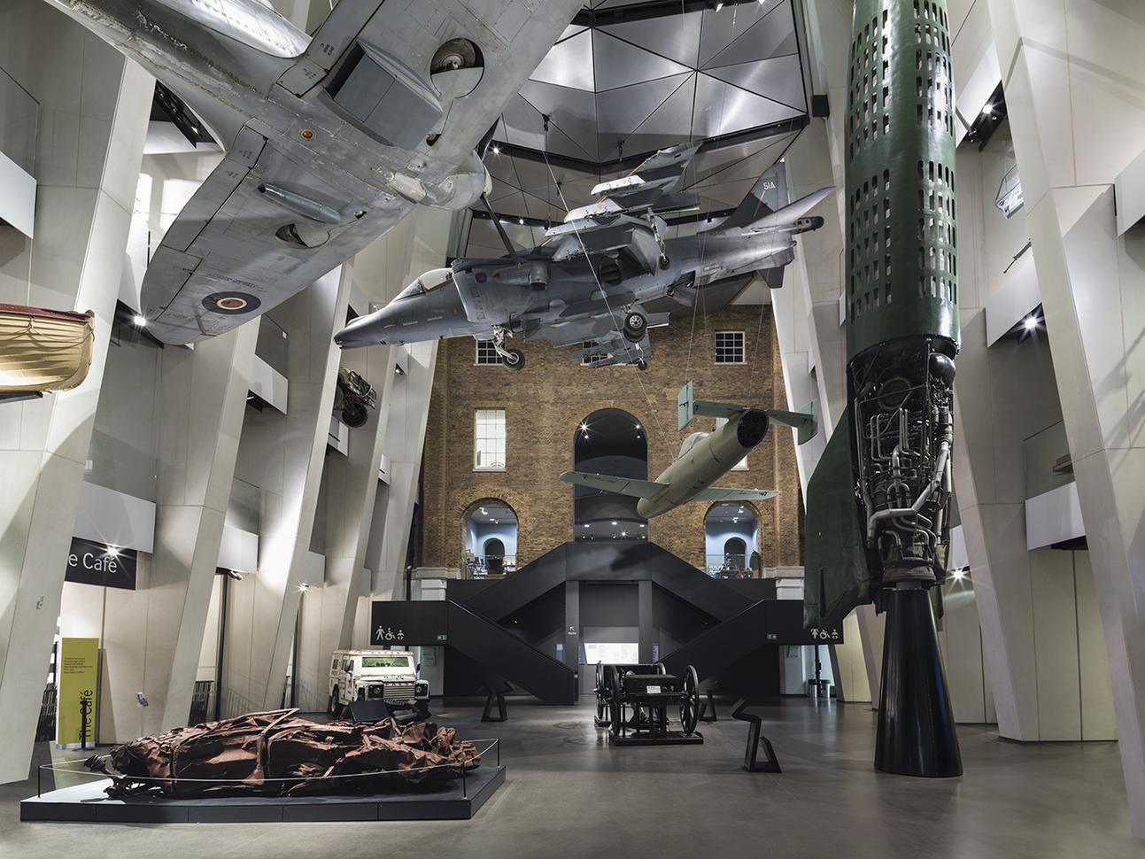 Photo of inside IWM London. The flooring has various images throughout the photo. There are planes located from the ceiling. There are stairs in the photo to the next level up.