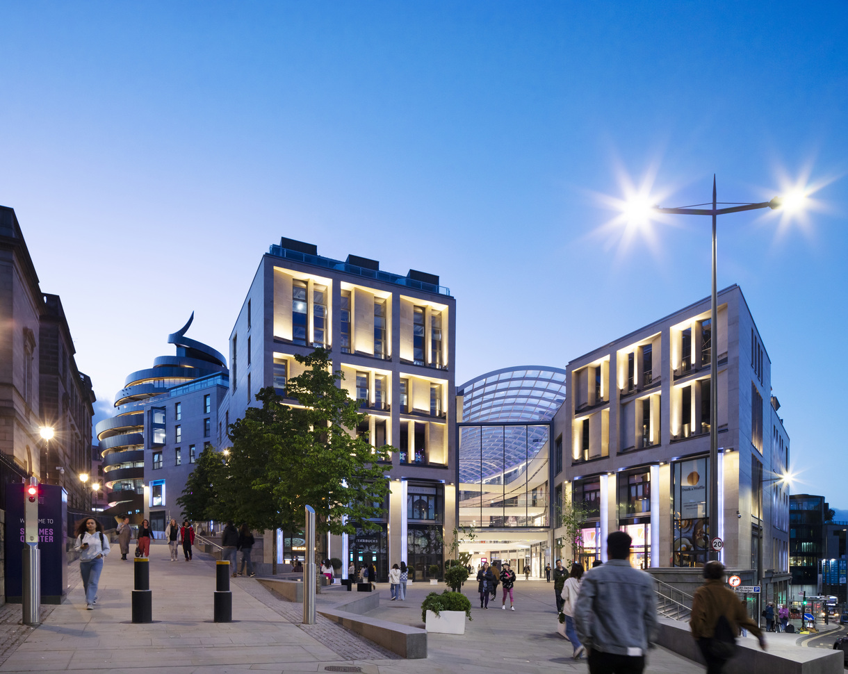 Modern city plaza at dusk with illuminated contemporary buildings, a glass-roofed entrance, and people walking along a wide pedestrian street under bright streetlights.