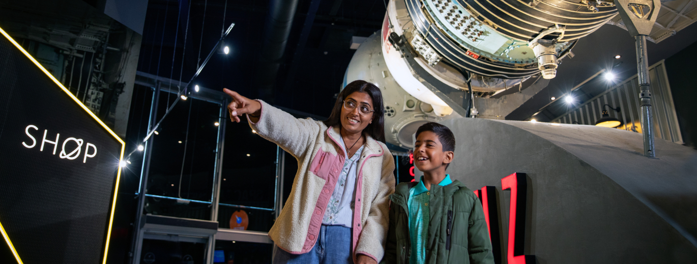 An Asian, lady with long dark hair wearing glasses and a pink jacket is stood in the National Space Centre, she is pointing to something in the distance, to her right is a young Asian boy  smiling wearing a green jacket, they are both standing in front of a rocket made for space exloration 