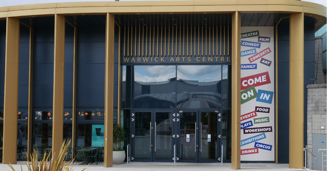 "Photo of main entrance to Warwick Arts Centre. There are gold/brown pillars either side of the entrance. There are 2 sets of double doors. There is a graphic on the right side of the entrance with words including theatre, workshops, drinks, come on in."