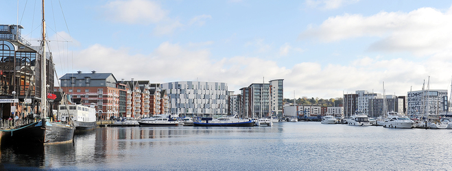 Waterfront campus setting with a marina, boats, and modern buildings along the shoreline under a bright sky