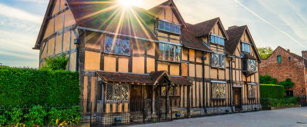 Large timber-framed Tudor-style building with exposed wooden beams and a tiled roof, lit by sunlight with a starburst effect, set along a hedge-lined street with a brick building nearby.
