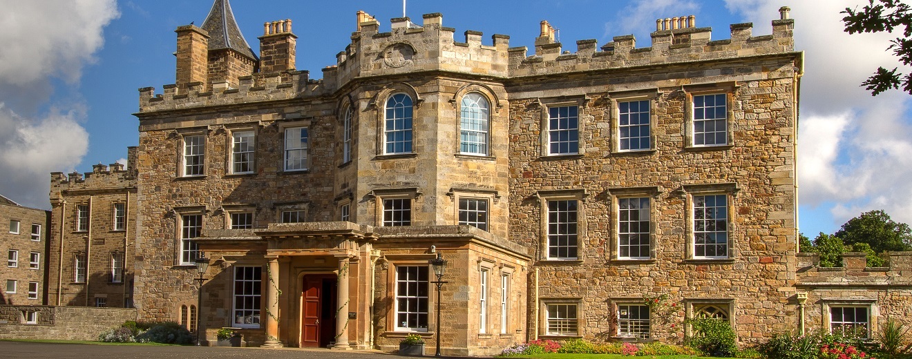 "Photo of the front entrance to Newbattle Abbey College. The building is all brick with numerous windows throughout. The entrance door is brown in the centre of the building with pillars either side."