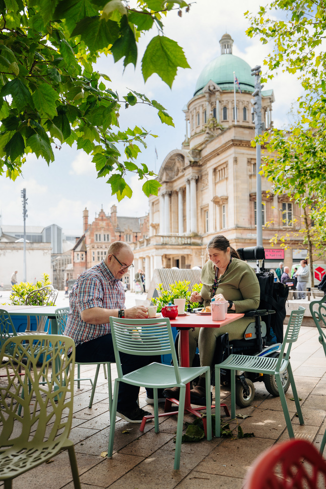 A man and woman sit at a tabl outside, eating tea and cake, the man is sat at the table on a chair, and the woman is in an electric wheelchair