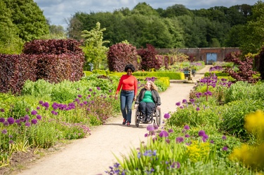 A person pushes a wheelchair user along a wide path through a colourful garden filled with purple flowers and neatly trimmed shrubs.