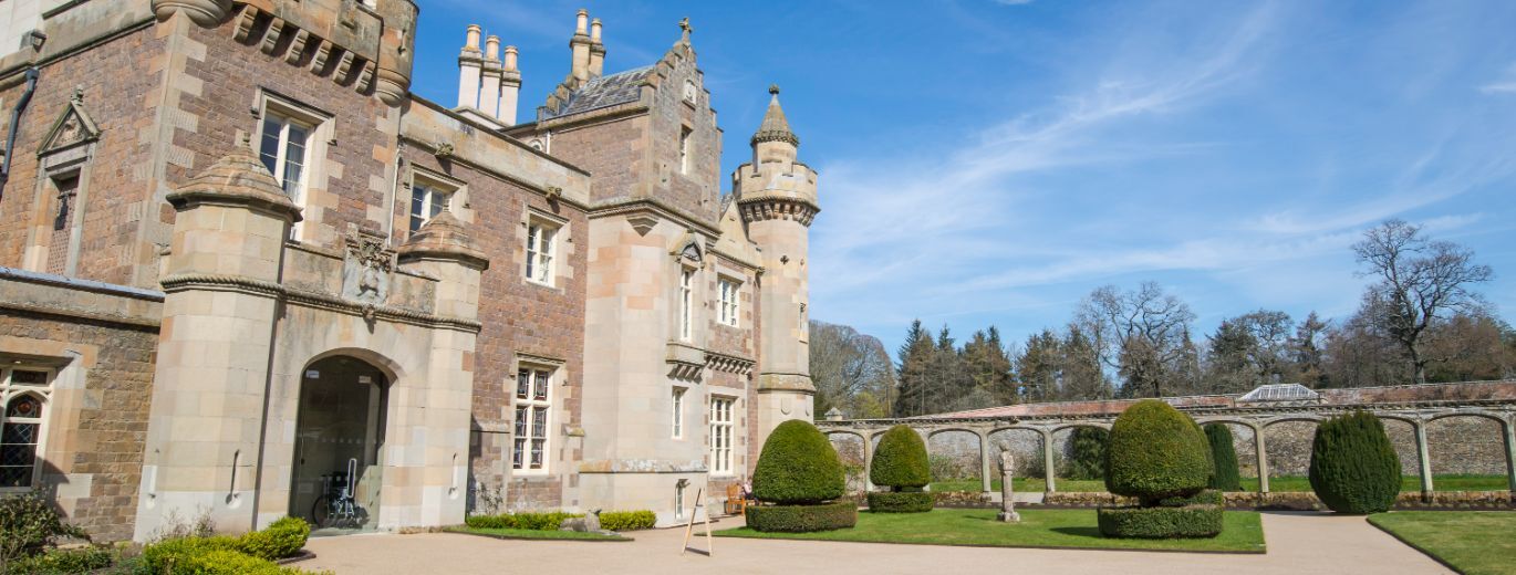"View of the entrance to Abbotsford House. A beautifully kept garden features either side of the a path leading to the entrance. The house is made of stone, looking almost like a castle with turrets and chimneys on top"