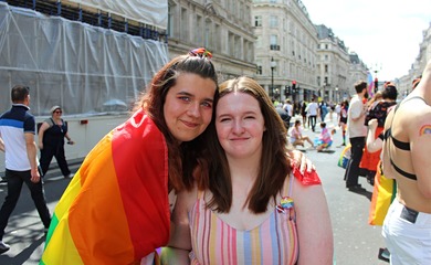 Jemma, a white non-binary person with long brown hair, and Shona, a white woman using a wheelchair, take a photo together at London Pride. They have their arms around each other and are draped in a rainbow pride flag. They are both smiling.