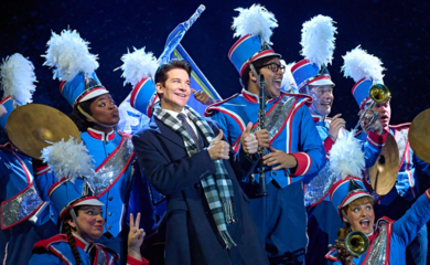 The Groundhog Day cast on stage at the Old Vic theatre. The main character is looking to the side with his thumbs up, as if he's posing for a photograph, surrounded by people dressed in marching band uniforms.