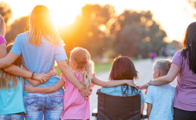 A diverse group of people wearing different coloured t-shirts, stood with their arms around each other watching the sunset.