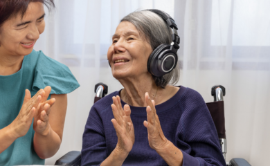 a woman stood next to her older relative, who is a wheelchair user and wearing noise-cancelling headphones. both are smiling and laughing together.