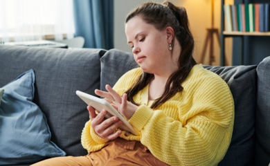 A young woman with a learning disability sat comfortably on the sofa, wearing a yellow jumper and looking down as she scrolls on the digital tablet in her hand.