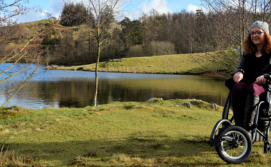 Carrie-Ann, a white female, using her manual wheelchair with a power add-on outdoors. She's wearing a coat and hat, with beautiful countryside lakes and hills visible behind her.