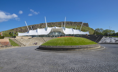 Outside of the Dynamic Earth attraction in Scotland, a futuristic-looking building situated at the top of a hill of grass with stepped and sloped access.