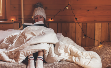 A young woman sitting on her bed and reading, looking cosy with blankets and warm socks. There are fairy lights and Christmas decorations on the walls.