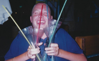 Ross is a white man and is pictured sat in his power chair. He wears a blue shirt with light coloured trousers. He is holding three large glasses with funnel necks, one containing a red alcoholic drink and the other two containing blue drinks. Each has a long straw in it. Ross is smiling to the camera from behind the drinks.