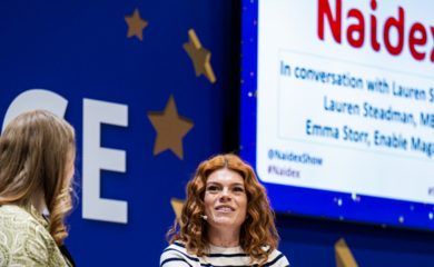 A photo from a previous Naidex event. In the background, you can see the word Naidex written. Two white women are facing each other. The one nearest the camera we see from the back. She has long, reddish brown hair. Opoosite her is a female with curly, reddish brown hair and she is wearing a black and white horizontally striped top.
