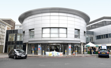Hospital entrance to 'North Middlesex University Hospital,' a circular modern building with a few cars parked nearby.