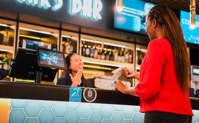 A black woman with long dark hair, wearing a red top and black trousers, stands at a bar in one hand; she has a receipt as the bartender signs ‘Thank You.’