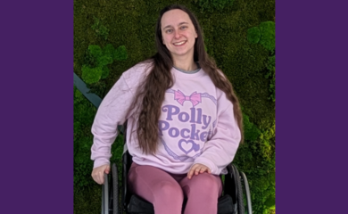 Katie Anna - a young white woman with brown wavy hair - sits in her old black wheelchair against a background of green shrubbery. She is smiling and wearing a pink Polly Pocket jumper to match her rose coloured leggings and sparkly silver trainers.